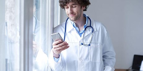 Ein junger Arzt lehnt am Fenster und schaut auf sein Handy. 