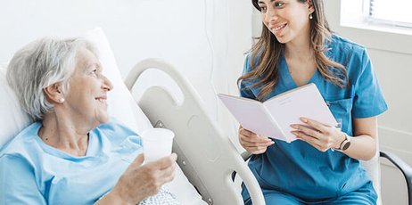 Eine ältere Patientin liegt im Krankenbett, eine junge Ärztin sitzt neben ihr und lächelt sie an. 