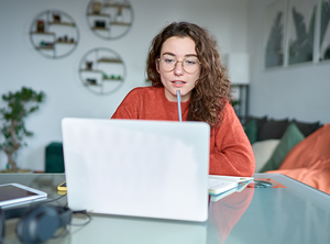 Eine junge Studentin sitzt zu Hause an ihrem Schreibtisch, schaut auf ihren Laptop und lernt.