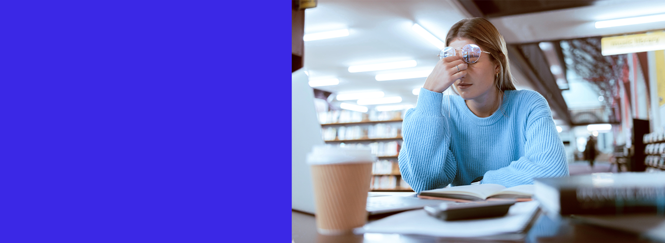 Eine Studentin sitzt in einer Bibliothek, vor ihr auf dem Tisch steht ein Laptop und liegen Arbeitsmaterialien. Sie reibt sich die Augen und kämpft mit Erschöpfung.
