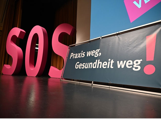Das Bild zeigt ein Plakat mit der Aufschrift „Praxis weg, Gesundheit weg.“ Im Hintergrund sind die Buchstaben „SOS“ zu sehen. 
