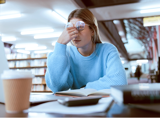 Eine Studentin sitzt in einer Bibliothek, vor ihr auf dem Tisch steht ein Laptop und liegen Arbeitsmaterialien. Sie reibt sich die Augen und kämpft mit Erschöpfung.