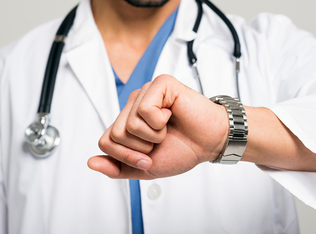 Arztzeit Artikelbild, indem man einen Oberkörper eines Arztes sieht, der mit einem weißen Kittel bekleidet ist und seinen linken Arm angewinkelt hat und auf die Uhr blickt.