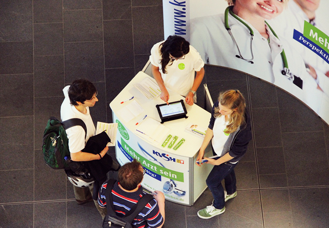 Vier Personen an einem Infostand mit Informationsmaterial, im Hintergrund ein großes Plakat