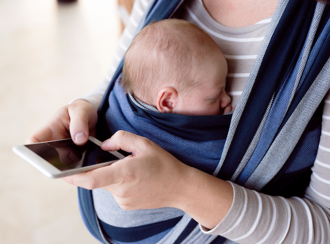 Mutter mit Kind in einem Baby Tragetuch und mit ihren Händen am Smartphone