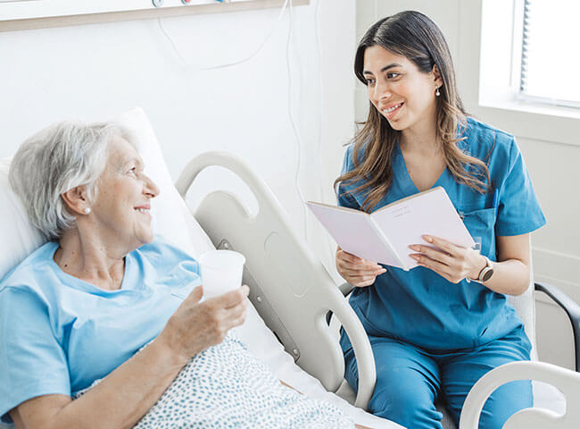 Eine ältere Patientin liegt im Krankenbett, eine junge Ärztin sitzt neben ihr und lächelt sie an. 