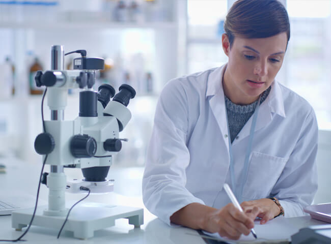 Eine Frau sitzt im Labor neben einem Mikroskop und macht Notizen für ihre Doktorarbeit. 