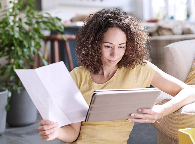 Eine Frau sitzt in ihrer Wohnung und prüft Dokumente auf ihrem Tablet.